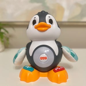 以下が含まれることがあります： 黒と白のボディ、オレンジ色の足とくちばし、ターコイズと白の翼を持つフィッシャー・プライスのペンギンの玩具。おもちゃには、光るセンターとベースのボタンがあります。フィッシャー・プライスのロゴが見えます。