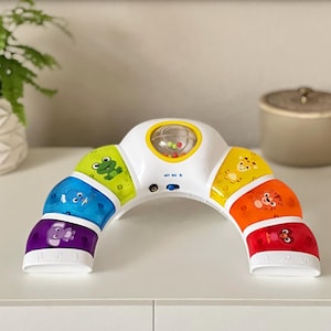 May include: A white and colorful baby toy in an arch shape. The toy features a clear rattle, and segments in green, blue, purple, yellow, orange, and red. Each segment has a different animal graphic. The text "en es fr" is printed on the toy.