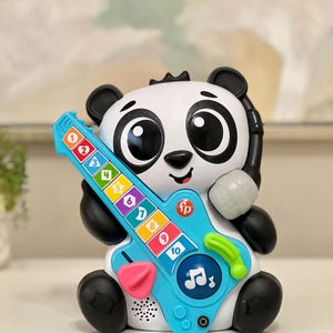 May include: A white and black panda toy with a blue guitar. The guitar has colorful buttons numbered 1-10. The toy has a microphone and the letters "fp" on the guitar. The panda has large black eyes and ears.