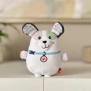 May include: A white plush dog toy with a blue collar and a musical note pendant. The dog has black and white patterned ears, a pink nose, and green and pink accents. The toy has a red tag.