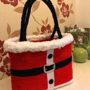 May include: Red and white crocheted Santa Claus bag with black handles and a black belt. The bag has white fur trim and black buttons.