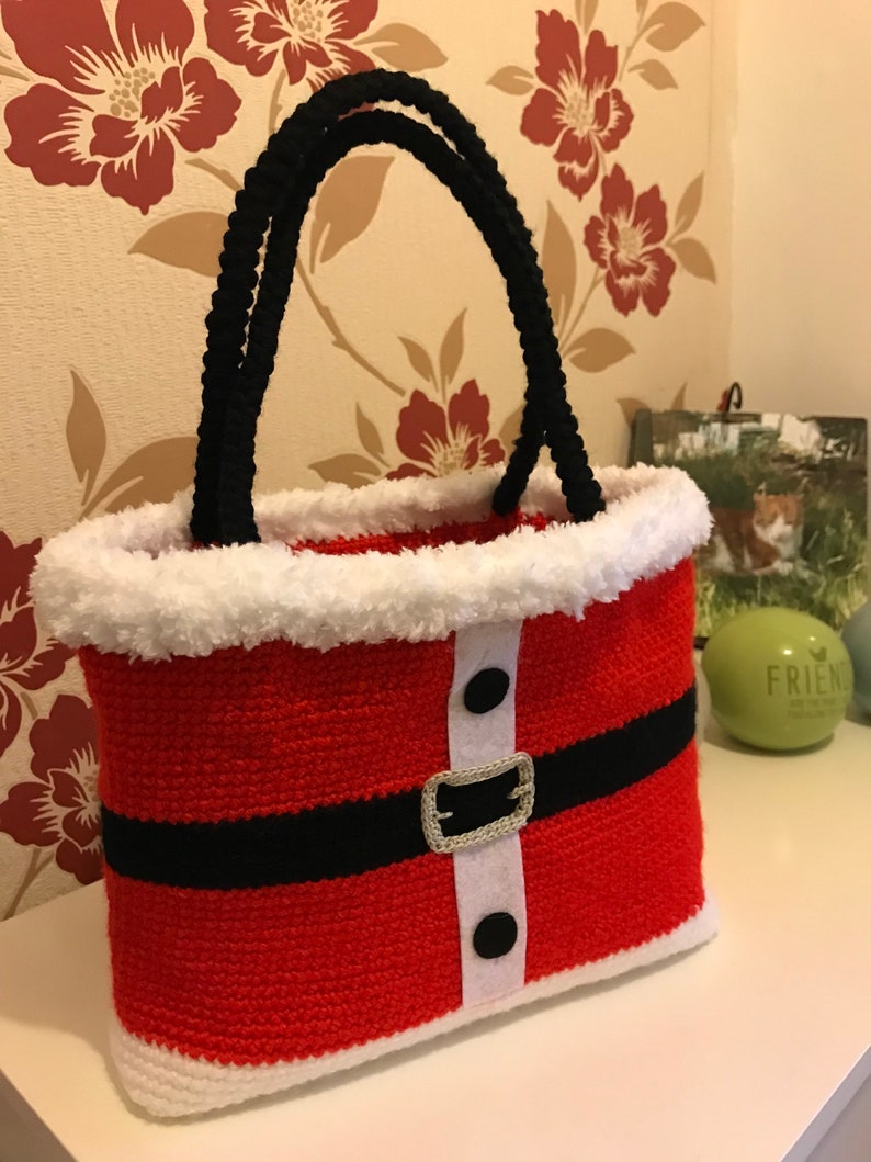 May include: Red and white crocheted Santa Claus bag with black handles and a black belt. The bag has white fur trim and black buttons.