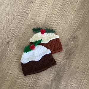 May include: Two crocheted Christmas pudding hats with white and brown yarn, green holly leaves, and red berries.