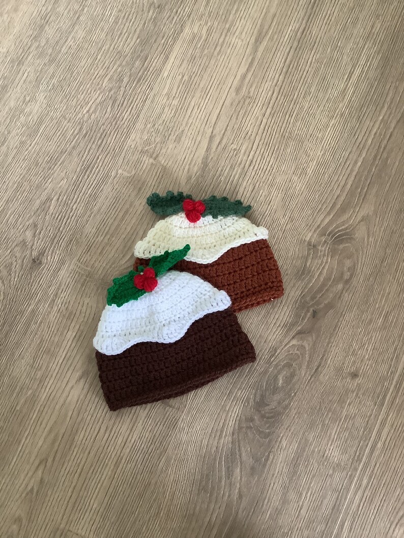 May include: Two crocheted Christmas pudding hats with white and brown yarn, green holly leaves, and red berries.