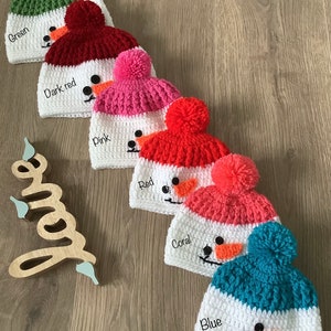 May include: Five crocheted snowman hats in different colors: green, dark red, pink, red, and blue. Each hat has a white brim and a white pom-pom on top. The hats are arranged in a row on a wooden surface.