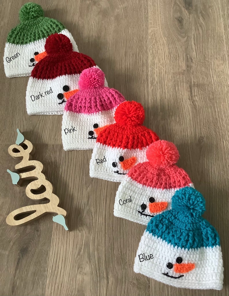 May include: Five crocheted snowman hats in different colors: green, dark red, pink, red, and blue. Each hat has a white brim and a white pom-pom on top. The hats are arranged in a row on a wooden surface.