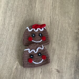 May include: Two brown crocheted gingerbread man hats with white icing and red buttons for eyes and cheeks. The top hat has a red bow.