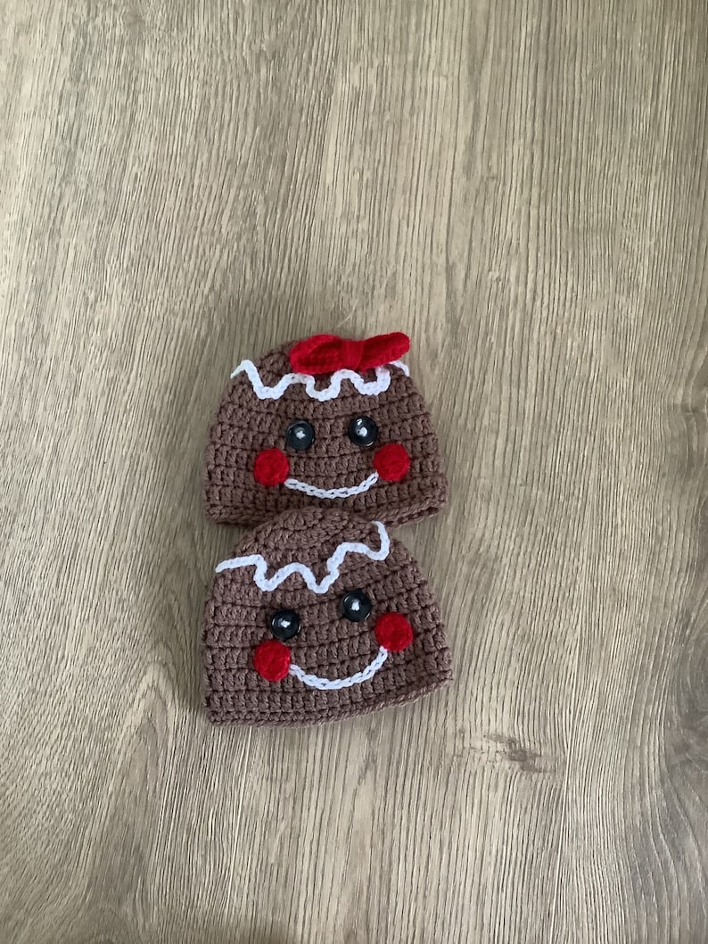 May include: Two brown crocheted gingerbread man hats with white icing and red buttons for eyes and cheeks. The top hat has a red bow.