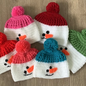 May include: Five crocheted snowman hats in different colors, including red, pink, green, blue, and white. Each hat has a white pom-pom on top and a snowman face with black eyes, a carrot nose, and a black smile.