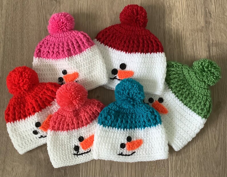 May include: Five crocheted snowman hats in different colors, including red, pink, green, blue, and white. Each hat has a white pom-pom on top and a snowman face with black eyes, a carrot nose, and a black smile.