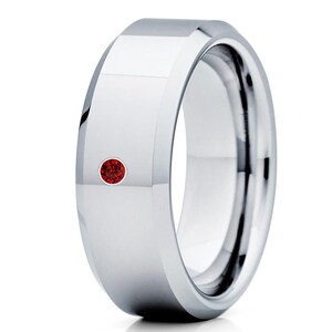 May include: A silver tungsten carbide ring with a beveled edge and a small red gemstone set in the center.