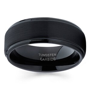 Puede incluir: Un anillo de carburo de tungsteno negro con acabado cepillado y borde biselado. El anillo tiene las palabras "Tungsten Carbide" grabadas en el interior.