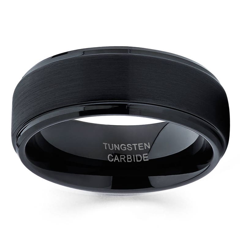 Puede incluir: Un anillo de carburo de tungsteno negro con acabado cepillado y borde biselado. El anillo tiene las palabras "Tungsten Carbide" grabadas en el interior.