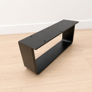 Könnte beinhalten: Ein mattschwarzes Möbelbein aus Metall mit rechteckigem, offenem Rahmendesign. Die obere horizontale Platte verfügt über vier vorgebohrte kreisförmige Löcher zur Montage, und die Struktur hat glatte, abgerundete Ecken an der Unterseite.