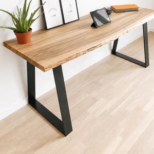 May include: A rectangular desk featuring a natural light-toned wood slab top with a live edge and two black metal trapezoidal legs. The desk surface holds a potted green aloe plant, two framed line art prints, a tablet, and a stack of notebooks.