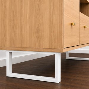 Op de afbeelding: Een kast van licht eikenhout met twee lades voorzien van kleine, goudkleurige rechthoekige handgrepen. De kast wordt ondersteund door een wit metalen frame met een rechthoekig ontwerp, wat een strak contrast vormt met de natuurlijke houtnerf.