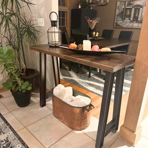 Op de afbeelding: Een rechthoekige houten sidetable met donkere metalen poten. Op het blad staan een zwarte draadstalen lantaarn, een zwarte decoratieve schaal met kleine kunstmatige pompoenen en een witte stompkaars. Daaronder staat een koperkleurige metalen bak gevuld met witte stof.