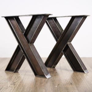Puede incluir: Dos patas de mesa de metal oscuro con diseño en forma de X. Las patas están hechas de acero grueso soldado y tienen una parte superior rectangular para fijar una mesa. Las patas están diseñadas para un aspecto robusto e industrial.