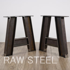 Op de afbeelding: Twee ruwe A-vormige tafelpoten van staal. De poten zijn gemaakt van zwaar staal en hebben een rustieke, industriële uitstraling. RAW STEEL is gedrukt op de houten vloer.