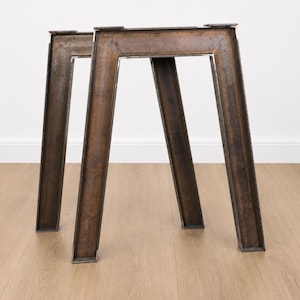Può includere: Due gambe per tavolo trapezoidali realizzate in metallo scuro e usurato con una patina visibile. Ogni gamba presenta un'ampia piastra di montaggio superiore e due supporti verticali angolati che si uniscono alla base, creando una struttura robusta.