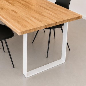 Könnte beinhalten: Ein rechteckiger Esstisch mit einer hellbraunen Holzoberfläche aus zusammengefügten Planken, getragen von einem dicken, weißen, U-förmigen Metallgestell. Zwei schwarze Stühle mit dünnen, dunklen Metallbeinen stehen um den Tisch herum.