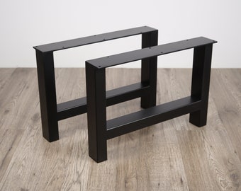 Könnte beinhalten: Zwei schwarze Metalltischbeine mit einem rechteckigen Rahmen und einer einzelnen Querstrebe. Die Beine stehen auf einem Holzboden.