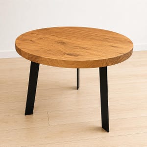 Könnte beinhalten: Ein runder Couchtisch mit einer Tischplatte aus naturfarbenem, hellbraunem Holz, das eine sichtbare Maserung, Astlöcher und Textur aufweist. Der Tisch steht auf drei stabilen, angewinkelten schwarzen Metallbeinen als Untergestell.