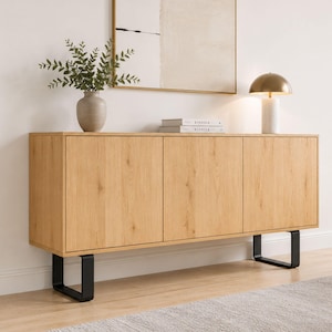 May include: A light oak wood sideboard with three doors and black metal legs. The top of the sideboard displays a vase with greenery, two books, and a gold and marble lamp. The sideboard is against a white wall.