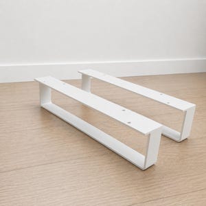 Op de afbeelding: Twee witte metalen meubelpoten met een rechthoekig U-vormig ontwerp. Elke poot heeft een platte bovenplaat met voorgeboorde ronde gaten voor montage. De poten hebben een gladde, matte afwerking en een strakke, lineaire structurele vorm.