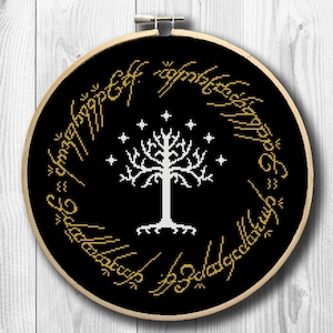 May include: A cross-stitch embroidery hoop featuring a black background with a white tree and stars design. Gold text encircles the design. The embroidery hoop is made of wood and is set against a white wooden background.
