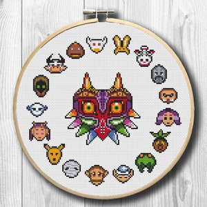 Puede incluir: Bordado de punto de cruz de Majora's Mask rodeado de máscaras de personajes del videojuego. El diseño está en un bastidor de bordado de madera, con fondo blanco y estilo pixelado.