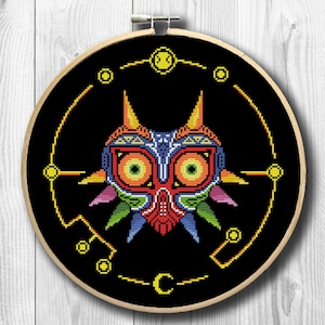 Op de afbeelding: Een kruissteekpatroon van Majora's Mask uit de The Legend of Zelda-videogameserie. Het masker is afgebeeld in een gepixelde stijl met een zwarte achtergrond en een gele cirkel eromheen. Het masker is rood, oranje, geel, groen, blauw en paars.