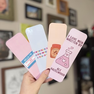May include: A hand holds four rectangular bookmarks. The bookmarks are pink, white, and peach-colored. Each bookmark has text and graphics, including a peanut butter jar, a beaker with hearts, and phrases like "I Love Men Written By" and "Hello I'm Antagonistic and Unapproachable."