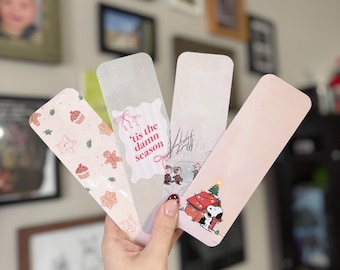 May include: Four Christmas-themed bookmarks are held in a hand. The bookmarks feature festive designs, including gingerbread men, Snoopy with a Christmas tree, and the text "'tis the damn season". The bookmarks have a rectangular shape.