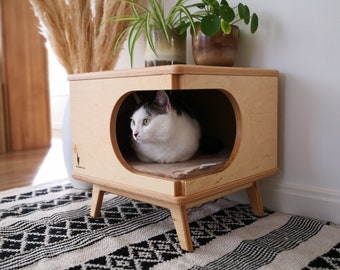 May include: A white and gray cat sits inside a wooden cat bed with a rounded opening. The bed is on a rug with a black and white geometric pattern. The bed has four legs and is made of light wood.