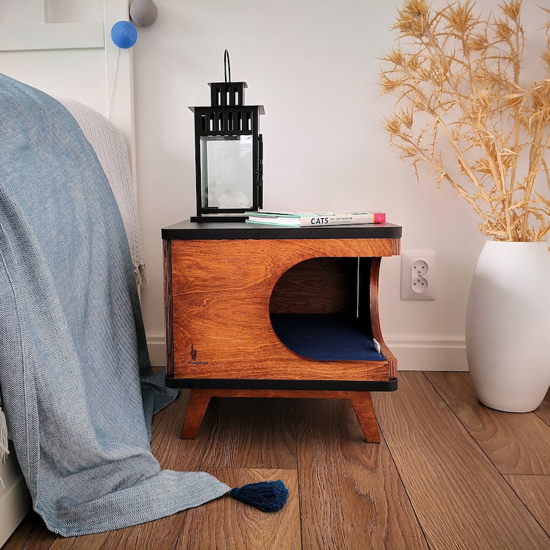 May include: A wooden cat bed with a black top and a blue cushion inside. The bed is on a wooden nightstand with a black lantern and a book titled "Cats" on top.