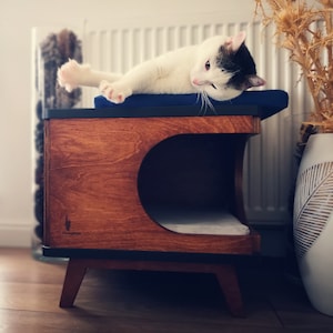 May include: A wooden cat bed with a black and white cat resting on a blue cushion. The bed has a rounded opening and a dark brown finish. The text "Plushtop" is engraved on the front of the bed.