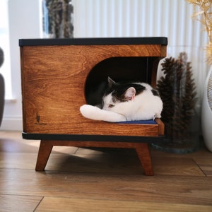 May include: A wooden cat bed with a black top and legs. The bed has a curved opening and a blue cushion inside. A black and white cat is sleeping on the cushion.