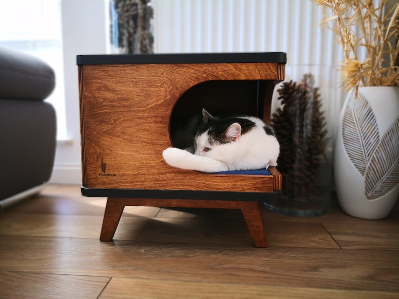 May include: A wooden cat bed with a black top and legs. The bed has a curved opening and a blue cushion inside. A black and white cat is sleeping on the cushion.