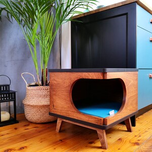 May include: A wooden pet bed with a blue cushion inside. The bed is shaped like a heart and has a dark brown finish. The bed is sitting on a wooden floor in front of a blue and black cabinet.