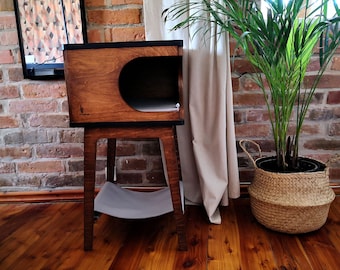 May include: A wooden cat bed with a curved opening and a shelf underneath. The bed is on a stand with a gray cushion on the shelf. The bed is in front of a brick wall and a plant in a wicker basket.