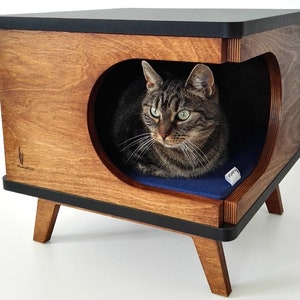 May include: A wooden cat bed with a black top and a blue cushion inside. The bed has a rounded opening and is on four legs. The cat bed is labeled "purrfect cat".