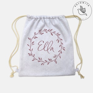 Puede incluir: Una bolsa con cordón de color gris claro con el nombre "Ella" en fuente cursiva, rodeada por una corona floral granate. La bolsa tiene cordones de color crema. Diseño sencillo y limpio, adecuado para transportar varios artículos.