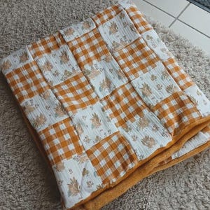 Peut inclure: Couverture patchwork pliée avec un motif de carreaux vichy orange et blanc et des panneaux à imprimé floral. La couverture a une bordure orange et repose sur un tapis de couleur neutre. La couverture est en tissu doux.