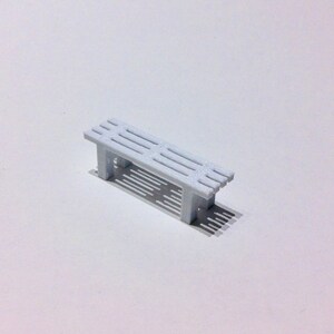 May include: A small, light blue bench with a slatted seat and four legs. The miniature bench casts a shadow on the white surface. This item is likely a decorative piece or part of a model.