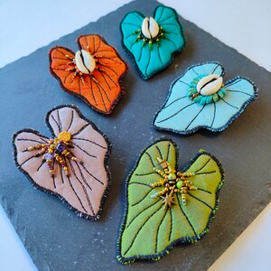 Puede incluir: Cinco broches de tela con forma de hoja con telas de diferentes colores y adornos de cuentas. Los broches se muestran sobre una superficie de pizarra gris oscuro.