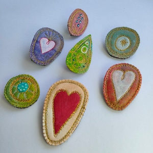 Könnte beinhalten: Sieben gestickte Filzpatches mit verschiedenen Designs, darunter Herzen, Sterne und Wirbel. Die Patches sind in den Farben Rosa, Grün, Weiß und Gold.