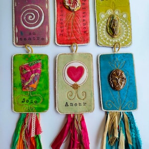 May include: Six handmade fabric ornaments with colourful fabric, embroidery, and embellishments. Each ornament has a different design and a French word written on it. The words are "Revenir au centre", "Intelligence et puissance", "Divine", "Joie", "Amour", and "Divine".