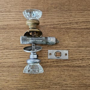 May include: An antique door knob set with a clear, faceted glass knob, brass collar, and silver-toned metal components. Includes a matching metal strike plate. The set is arranged on a wooden surface.