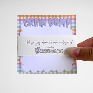Peut inclure: Un bloc-notes avec une bordure à carreaux colorés et les mots "BRAIN DUMP" en violet. Le bloc-notes est maintenu par une bande blanche qui indique "30 pages handmade notepad" et "made by JOUNE'S JOURNALS".
